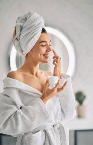 Xu Hướng Làm Đẹp Mới Nhất 2025: Công Nghệ Gặp Gỡ Tự Nhiên 2 Phụ nữ mặc áo choàng tắm trắng, quấn khăn trên đầu, đang thoa kem dưỡng da mặt và mỉm cười thư giãn trong phòng tắm hiện đại – xu hướng chăm sóc da tại nhà năm 2025.