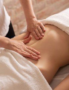 Nhân viên trị liệu đang thực hiện massage nhẹ nhàng trên vùng lưng dưới của một người đang nằm úp, được phủ khăn trắng. Không gian ấm cúng và sạch sẽ, mang lại cảm giác thư giãn, chăm sóc sức khỏe sau thẩm mỹ hoặc điều trị."